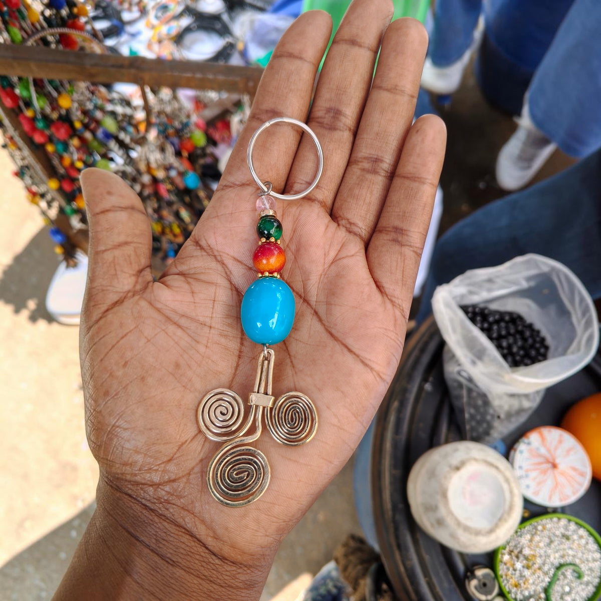 Nualocal handcrafted key holder with brass triple-spiral motif, blue resin bead, and colorful accents in a palm.
