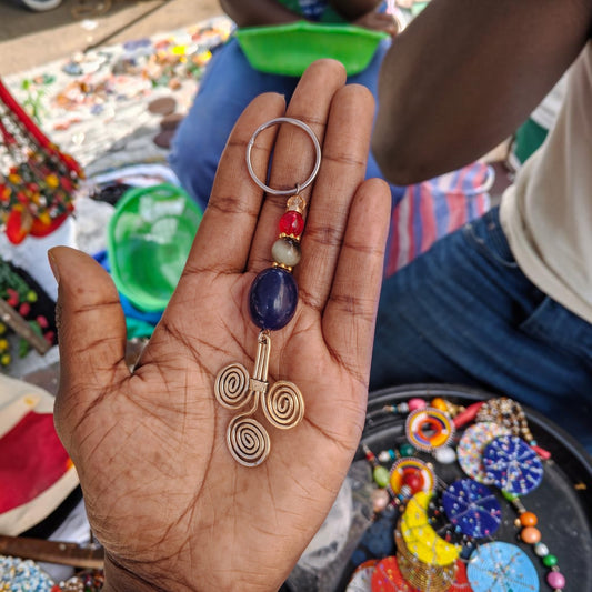 Nualocal handcrafted key holder featuring a brass triple-spiral motif with blue, grey, and red resin beads in a palm.