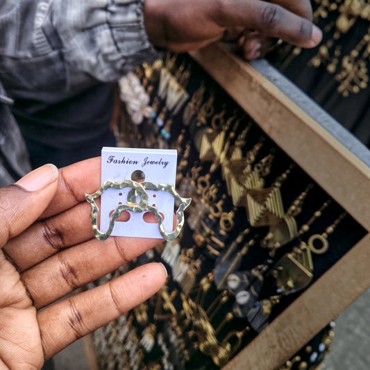 Nualocal handcrafted solid brass Africa map earrings with a hammered texture held in a hand at an outdoor market.