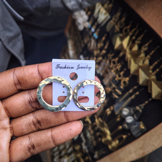 Nualocal hammered brass hoop earrings with an organic textured finish held in a hand against a jewelry display