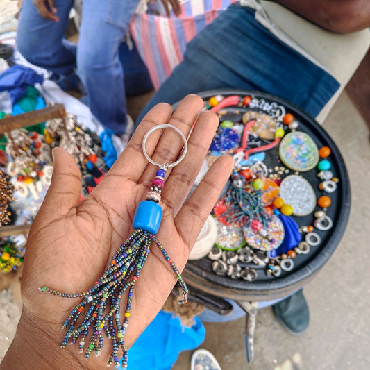 Nualocal handcrafted rainbow beaded tassel keychain with a blue focal bead and metal ring held in a hand.