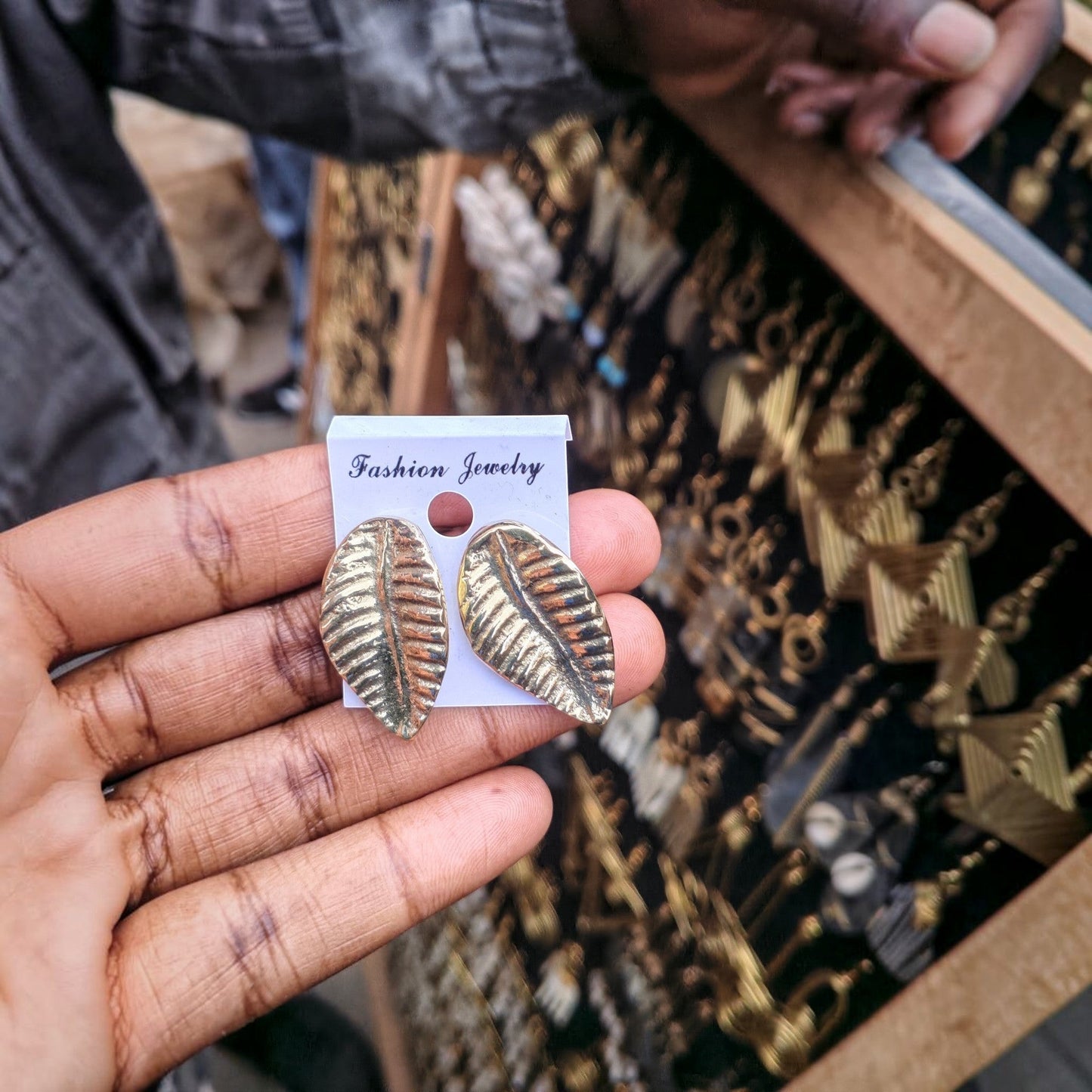 Nualocal textured leaf brass stud earrings with detailed veins held in a hand against a display of artisan jewelry.