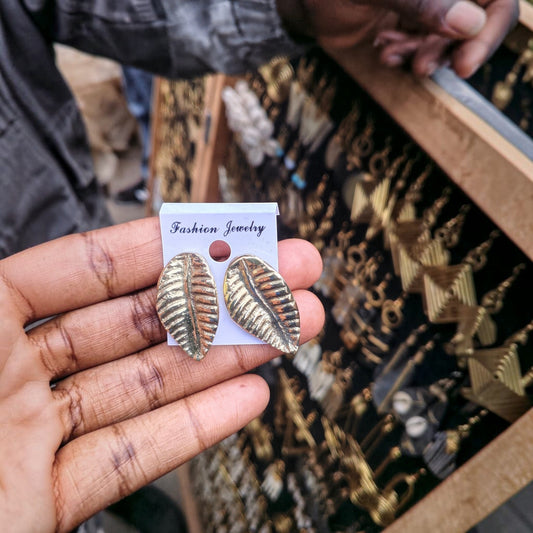 Nualocal textured leaf brass stud earrings with detailed veins held in a hand against a display of artisan jewelry.