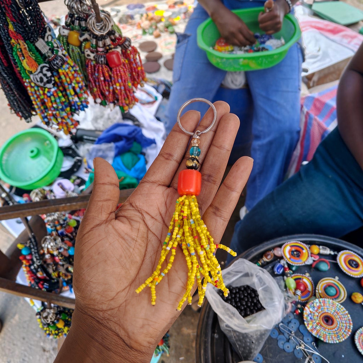 Nualocal handcrafted amber resin and sunshine yellow glass beaded tassel keychain held in a palm at a market stall.